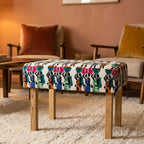 A square upholstered bench with a colorful patterned fabric top and light wood legs, placed in a warm living room setting with chairs and a rug in the background.