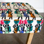 A square upholstered bench or ottoman with a colorful African-inspired print of figures carrying baskets, supported by two light wood legs on a concrete floor.