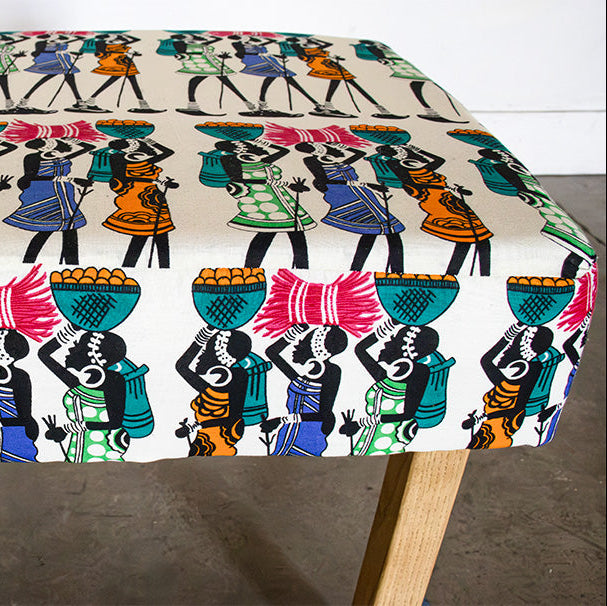 A square upholstered bench or ottoman with a colorful African-inspired print of figures carrying baskets, supported by two light wood legs on a concrete floor.