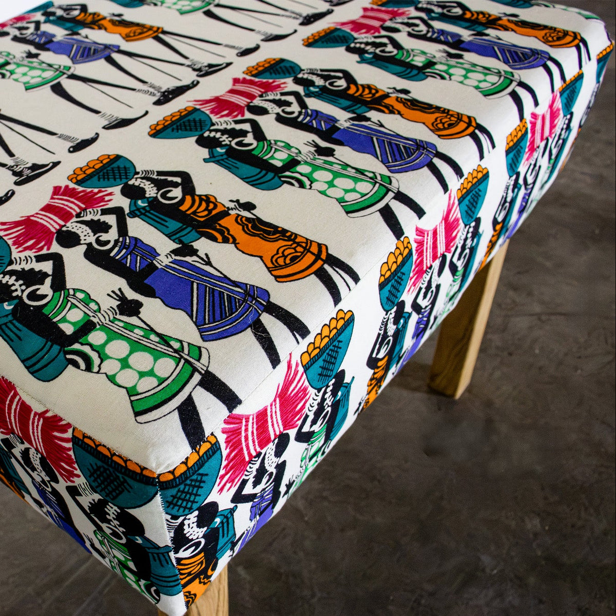 A rectangular upholstered bench with a colorful printed fabric featuring stylized figures in red, blue, green, orange, and black on a cream background, supported by light wood legs.