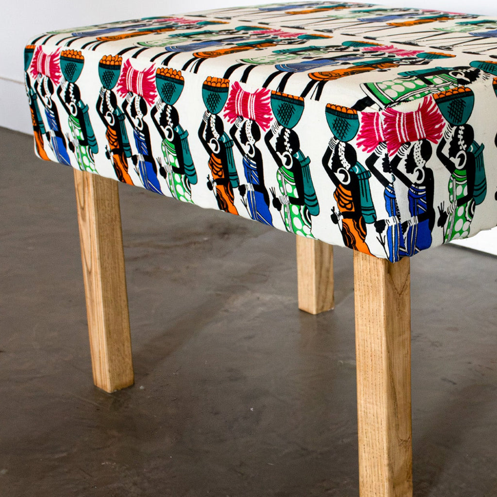 A square upholstered bench with a colorful, patterned fabric top and four natural wood legs, shown in a room setting with a concrete floor and white wall.