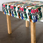 A square upholstered bench with a colorful, patterned fabric top and four natural wood legs, shown in a room setting with a concrete floor and white wall.