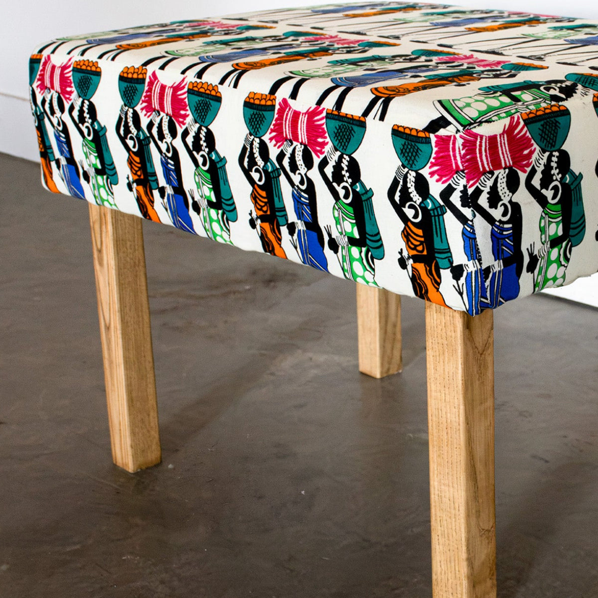 A square upholstered bench with a colorful, patterned fabric top and four natural wood legs, shown in a room setting with a concrete floor and white wall.