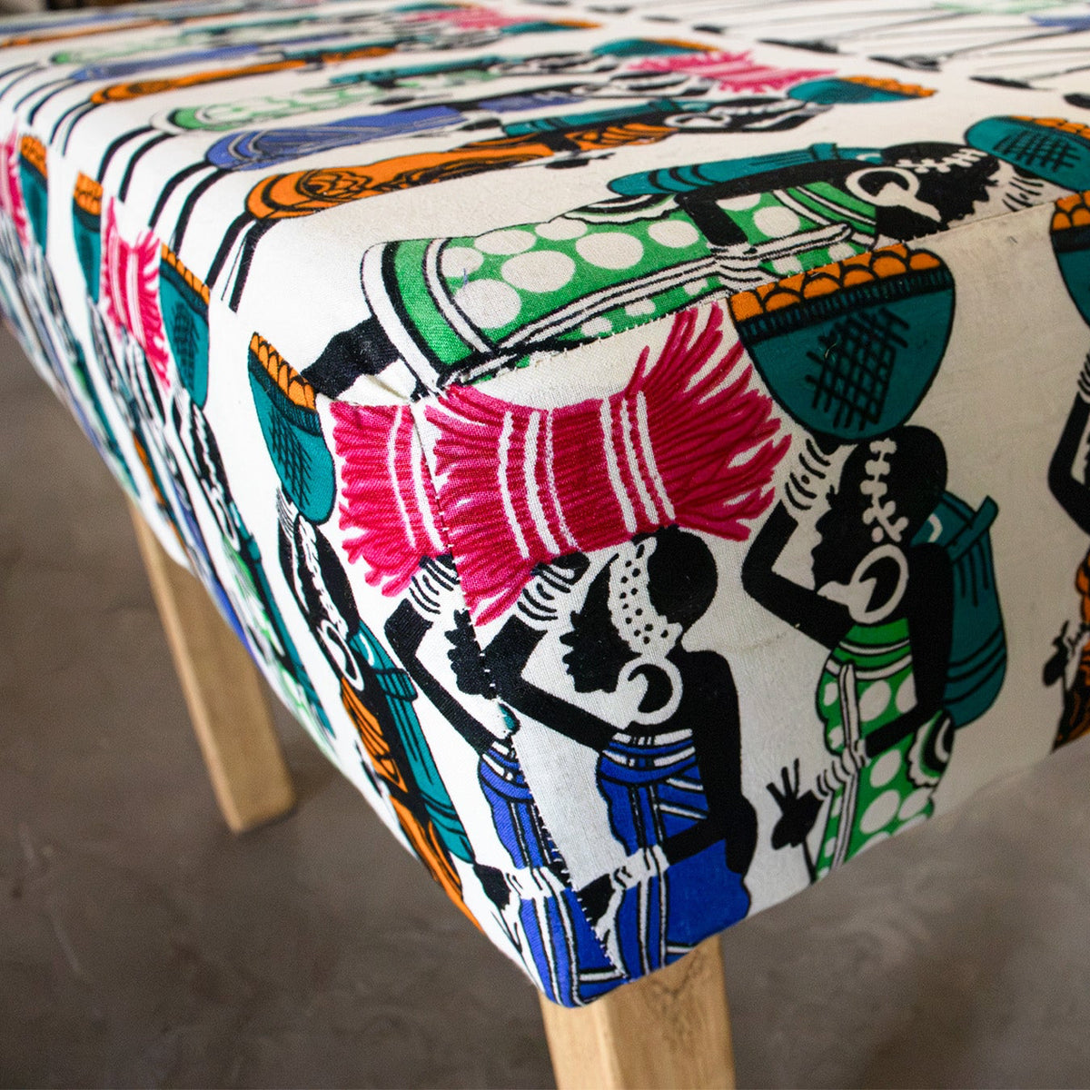 A close-up of a upholstered bench or ottoman with a colorful African-inspired print featuring stylized figures and baskets on a cream background, supported by light wood legs.